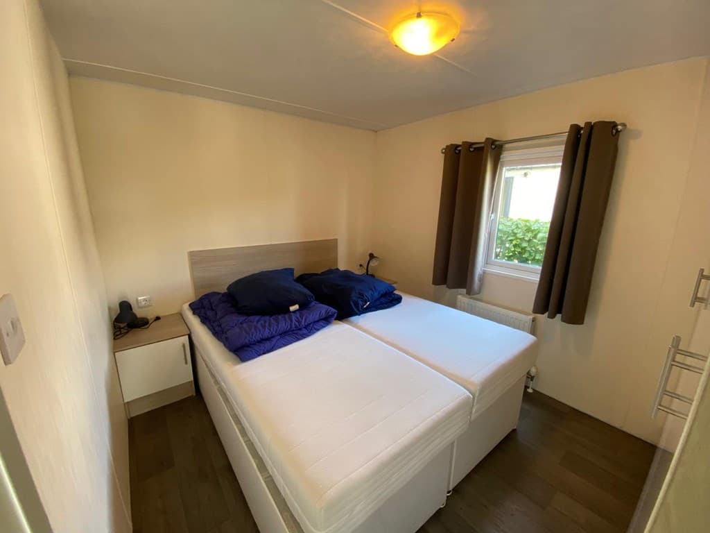 Slaapkamer met twee losse bedden, nachtkastje en gordijnen bij een raam. Houten vloer zichtbaar.