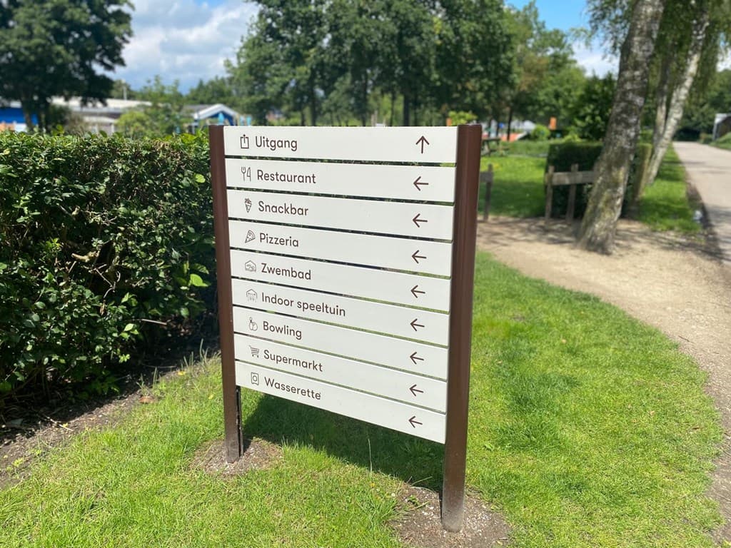 Informatiebord met verschillende locaties en richtingen in EuroParcs Zuiderzee, omgeven door bomen en gras.