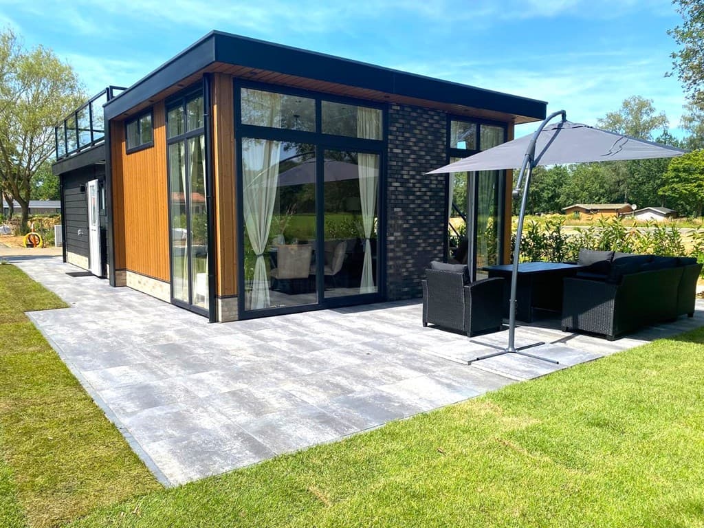 Moderne woning met grote ramen, terras met tuinstoelen en parasol, omgeven door gras en bomen.
