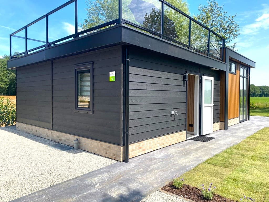 Moderne vakantiewoning met houten gevel, een veranda en een dakterras, omgeven door grasvelden en natuur.