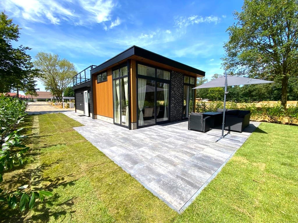 Moderne vakantiewoning met grote ramen, terras en zithoek, omgeven door groene tuin en bomen.