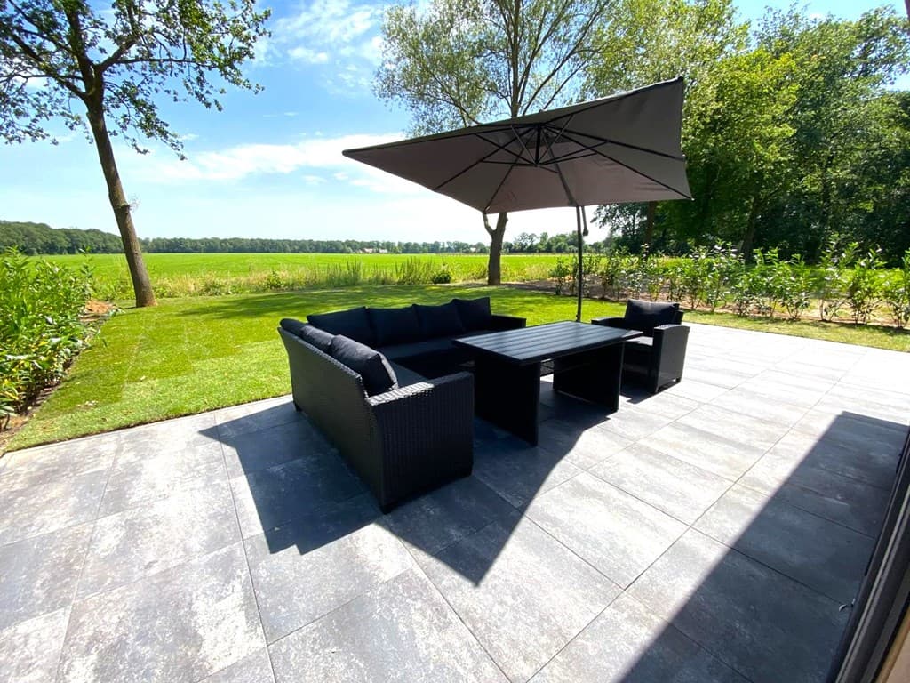 Terras met zithoek van wicker meubelen en parasol, omgeven door groene natuur en een grasveld.
