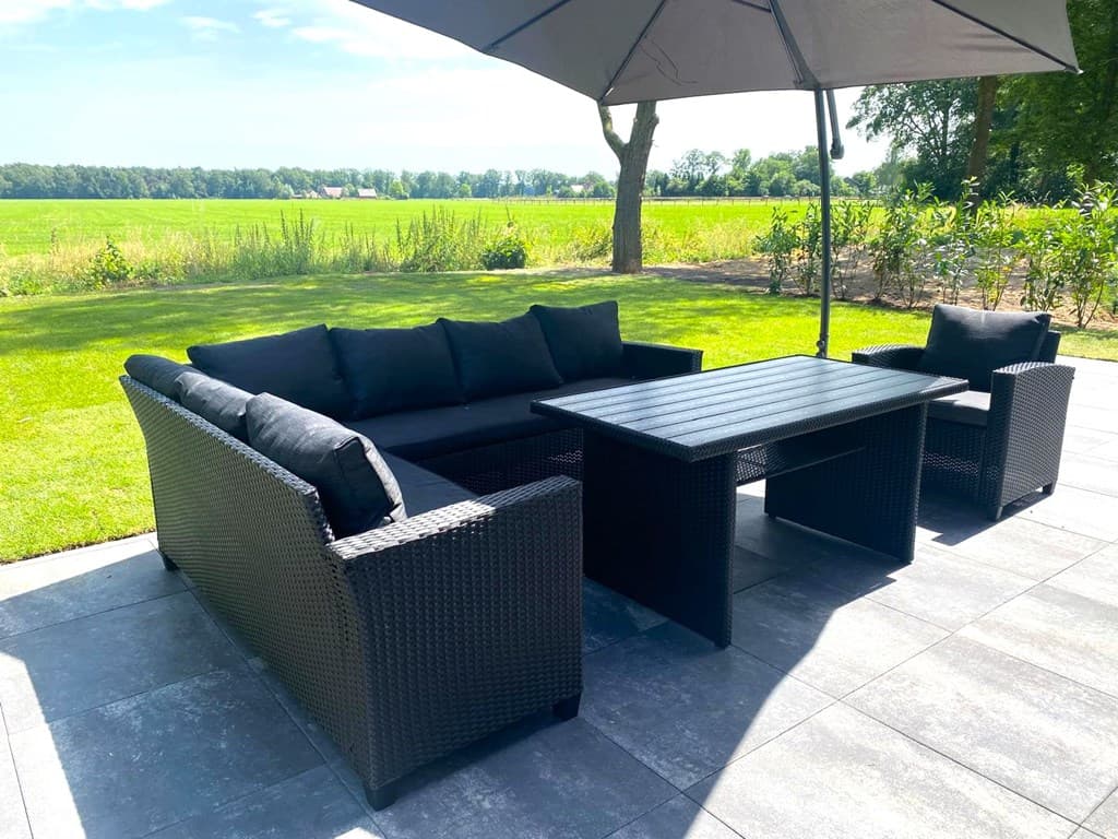 Tuin met een zitgedeelte bestaande uit een loungebank en een tafel, onder een parasol met uitzicht op groen landschap.