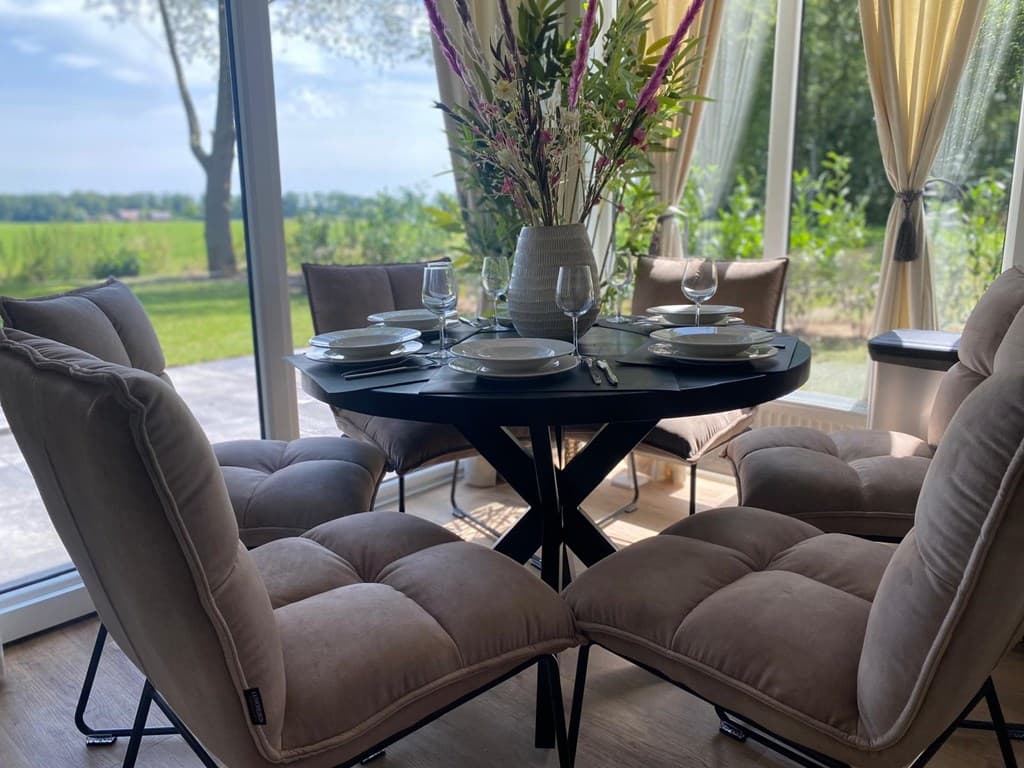 Eetgedeelte met donker ronde tafel, zes comfortabele stoelen en een vaas met bloemen bij grote ramen met uitzicht op de tuin.