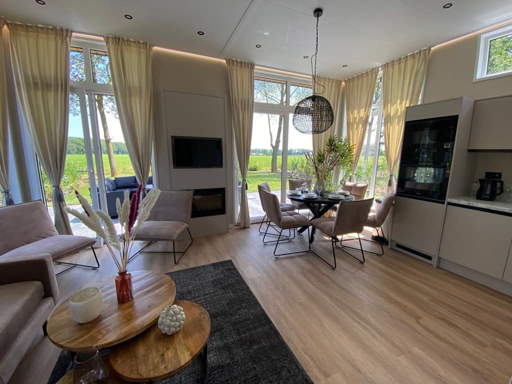 Moderne woonkamer met grote ramen, eetkamertafel en uitzicht op de tuin met groene bomen en veld.