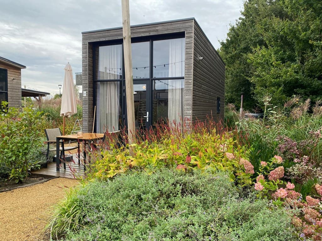 Moderne vakantiewoning omringd door groen en bloeiende planten, met een klein terras en een parasol.
