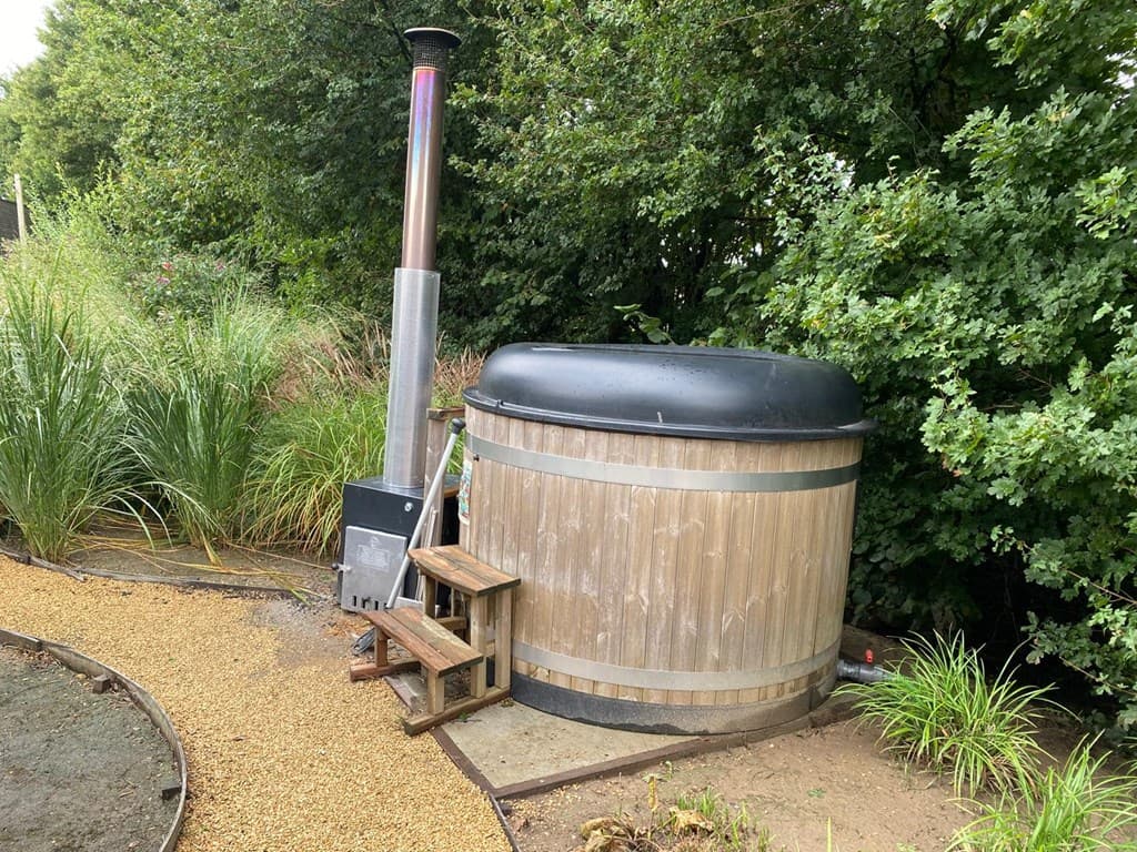 Houten hottub met een kachel, omringd door groen en een grindpad.