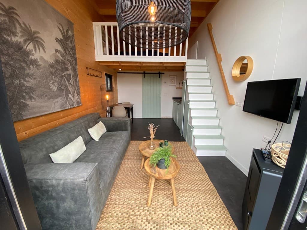 Zithoek met grijze bank, twee tafels, tv en trap naar de bovenverdieping in een moderne vakantiewoning.