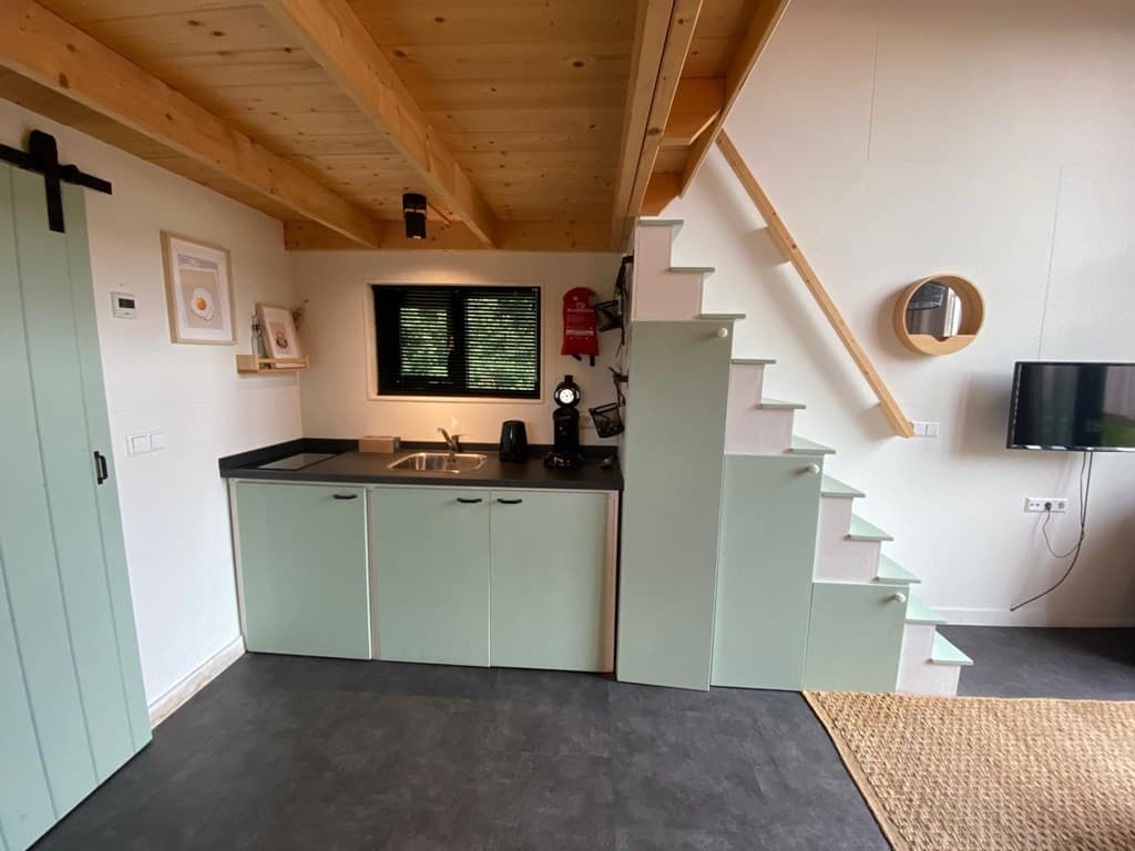 Gezellige woonkamer met keuken, houten plafonds en trap naar boven, meubels in lichte kleuren en een tv aan de muur