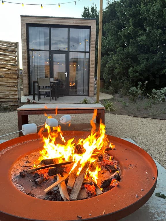 Gezellig kampvuur met brandende blokken en marshmallows, met uitzicht op een moderne woning in het vakantiepark.