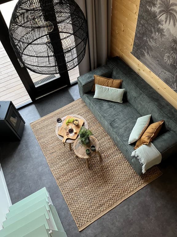 Zicht op de zithoek met groene bank, twee houten bijzettafels en een groot plafondlamp boven een jute vloerkleed
