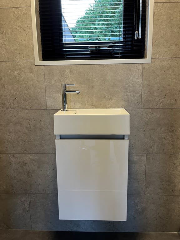 Witte wasbak met moderne kraan en meubel, tegen een tegelwand met een raam met jaloezieën.