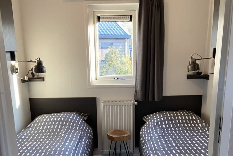 Slaapkamer met twee eenpersoonsbedden, een raam met gordijn en een houten krukje tussen de bedden.