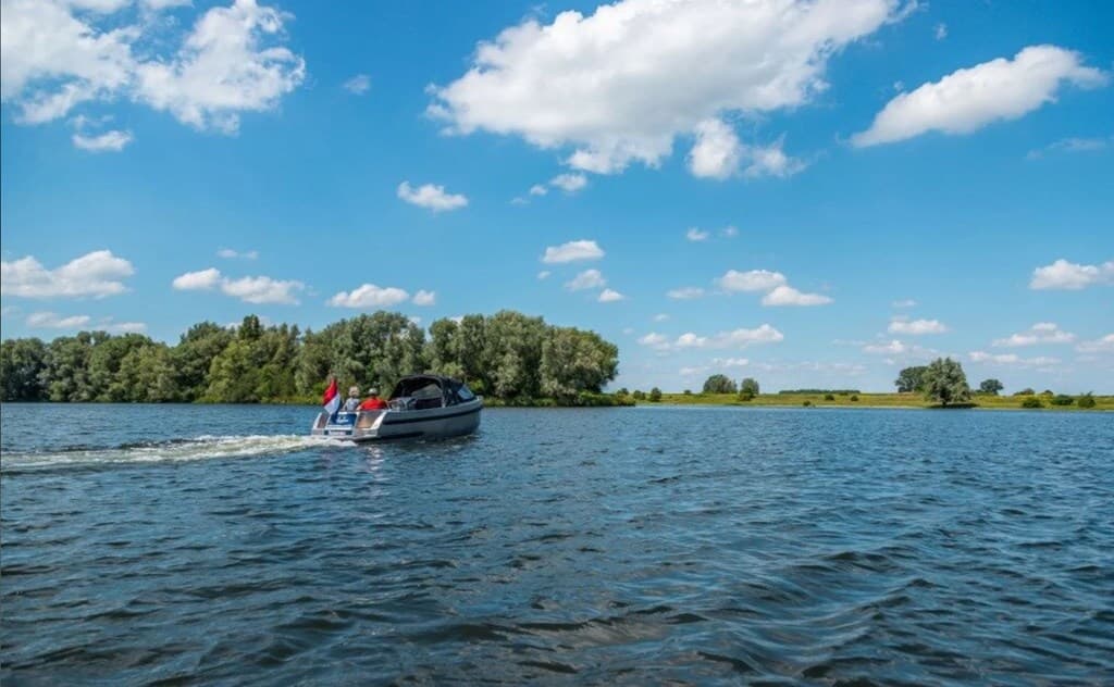 Een speedboot vaart over het water van de Maas met groene oevers en een heldere blauwe lucht op de achtergrond