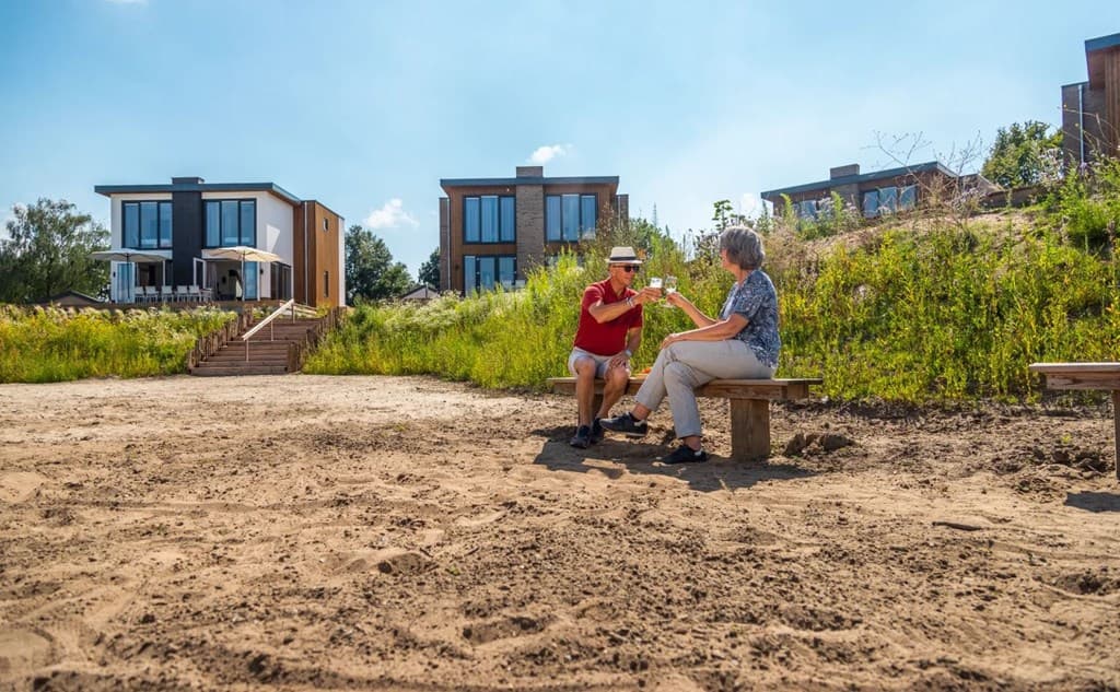 Zicht op vakantiehuisjes boven een zandstrand, met twee mensen op een bankje in het zand en groen rondom.