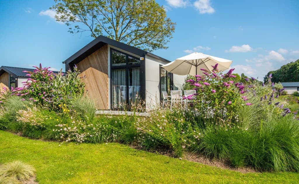 Moderne vakantiewoning met grote ramen, omringd door bloemen en groen, en een parasol op het terras