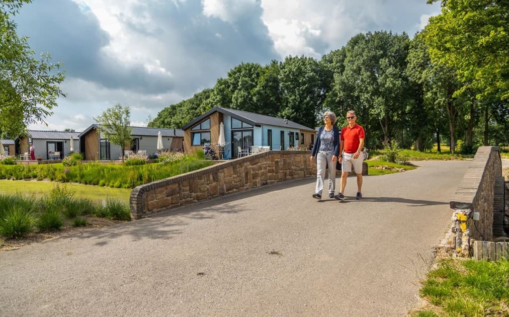 Twee mensen wandelen over een stenen brug met uitzicht op vakantiewoningen en groene bomen aan de Maas.