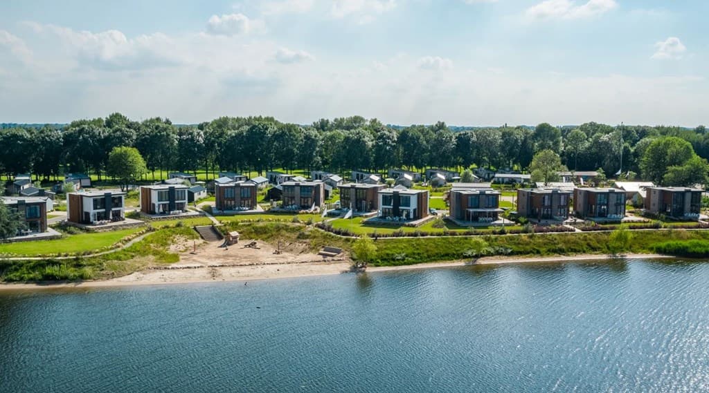 Uitzicht op vakantiewoningen aan de Maas met groene tuinen en zandstrand langs het water.