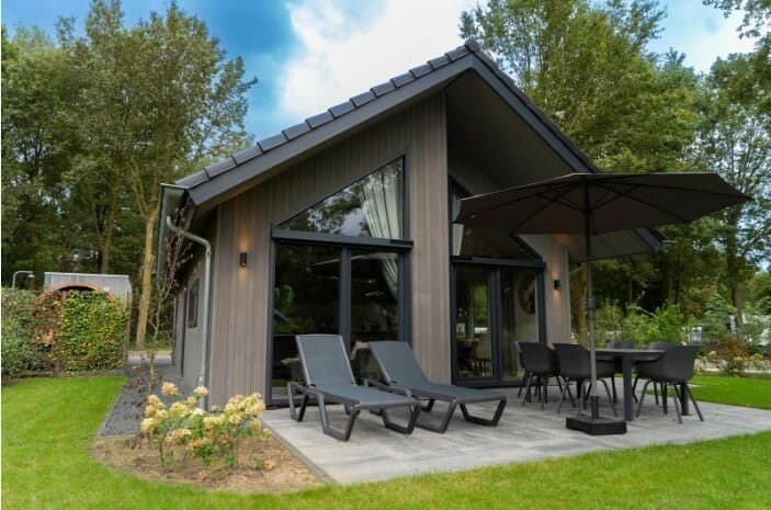 Moderne vakantiewoning met terras, ligstoelen en parasol, gelegen in groene omgeving met bomen en gras.