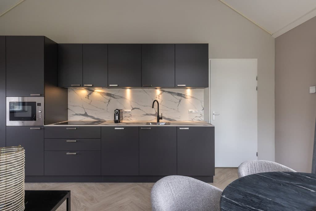 Moderne keuken met donkere kasten, marmeren achterwand, ingebouwde oven en zwarte kraan, vergezeld door een ronde tafel.