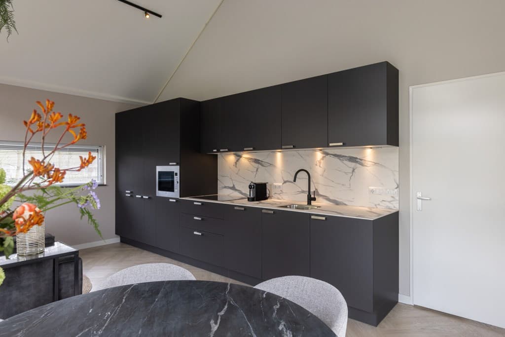 Moderne keuken met zwarte kasten, marmeren aanrecht en inbouwapparatuur, naast een ronde eettafel.