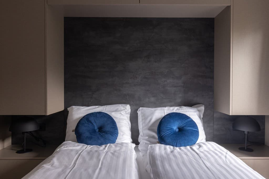Slaapkamer met twee eenpersoonsbedden en blauwe kussens tegen een grijze muur met ingebouwde verlichting.