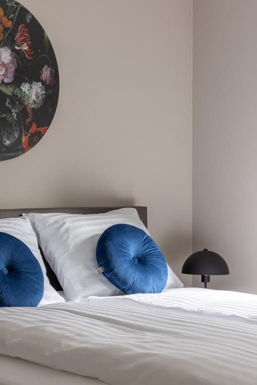 Slaapkamer met een bed, twee witte kussens en blauwe decoratieve kussens, een lamp en een kunstwerk aan de muur