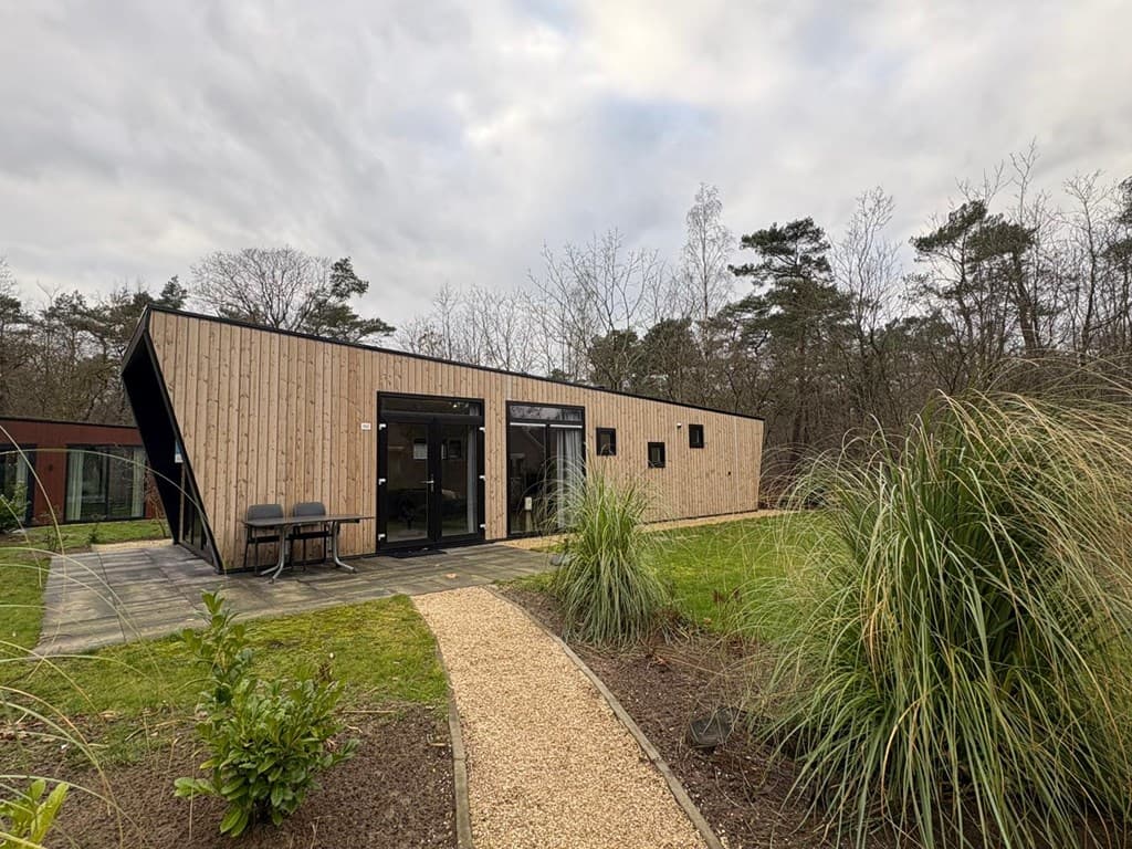 Moderne vakantiewoning met houten gevel, grote ramen en terrasje, omgeven door een groene tuin en paden van grind.