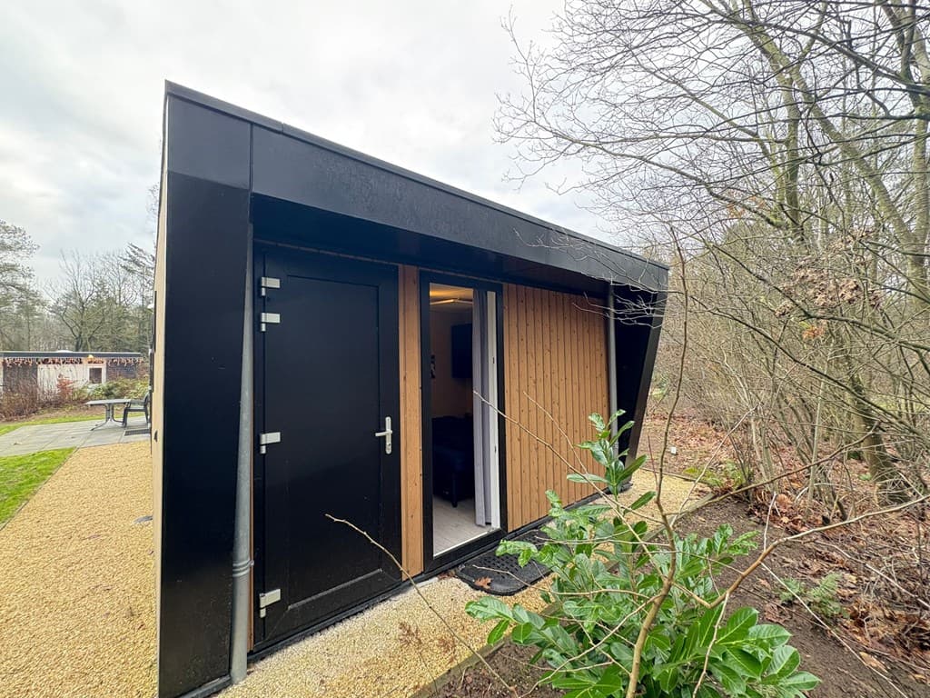 Moderne vakantiewoning met zwarte gevel en houten accenten, omgeven door bomen en planten, toegang via een zwarte deur