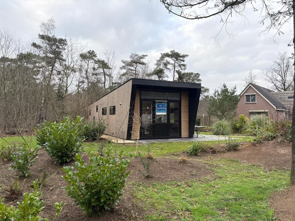 Moderne vakantiewoning met grote ramen en een entree, omgeven door tuinplanten en bomen in een bosrijke omgeving.