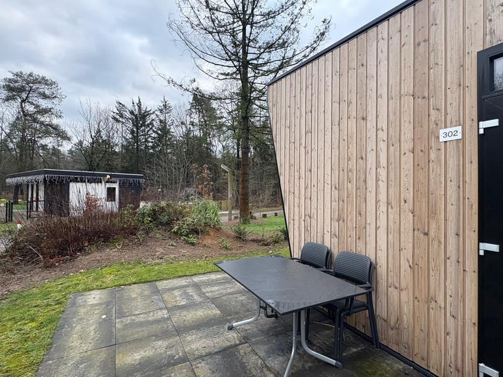 Houten wand van een vakantiewoning met een tafel en stoelen op een betegeld terras, bomen en bushaltes op de achtergrond