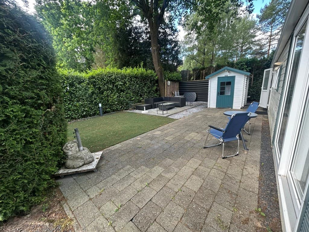Tuin met betegeld terras, grasveld, bank en stoelen, omringd door hoge hagen en een houten schuur.
