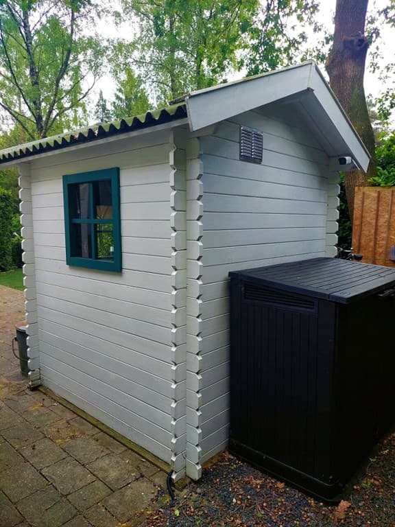 Houten berging met een zwart opslagbox aan de zijkant, omgeven door groene bomen en gras.