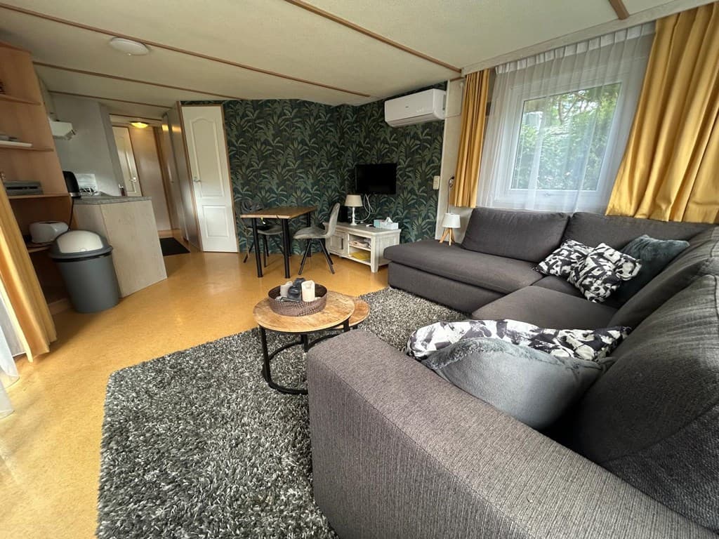 Ruime woonkamer met een grijze bank, eettafel, televisie en groene wanddecoratie in een vakantiehuis.