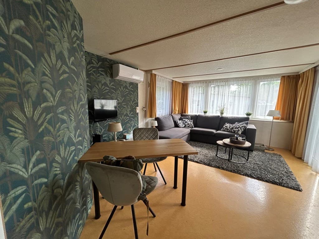 Gezellige woonkamer met groene wand, bank, eettafel en veel natuurlijk licht door grote ramen.