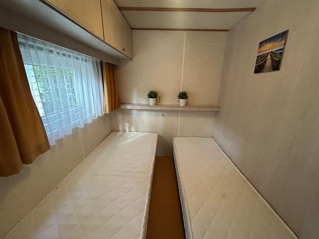 Slaapkamer met twee enkele bedden, een raam met witte gordijnen en een wandplank met planten op een neutrale achtergrond