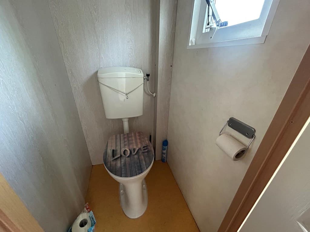Klein toilet met een houten toiletdeksel met de tekst 'LOVE', een raam en een toiletrolhouder aan de muur