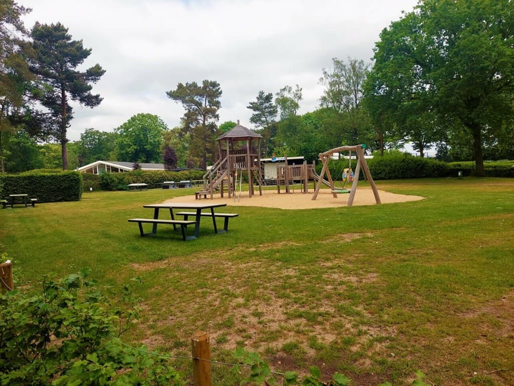 Speeltuin met houten speeltoestellen en schommels, omgeven door gras en bomen, met picknicktafels in de buurt.