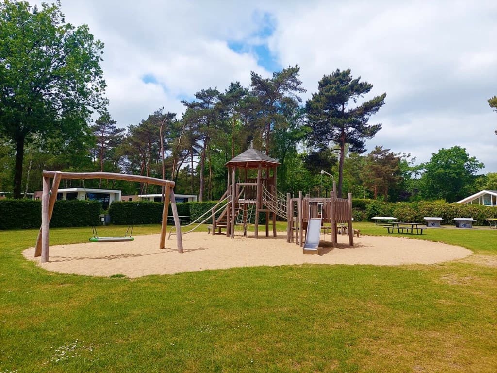 Speeltuin met zandgrond, houten speeltoestellen, schommel en picknicktafels omgeven door bomen en gras.