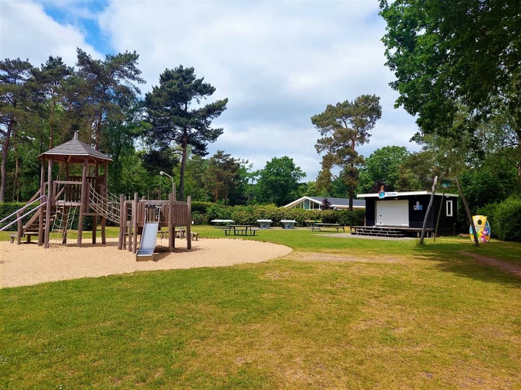 Speelplaats met houten speeltoestellen en een grasveld omgeven door bomen, met een recreatieruimte op de achtergrond