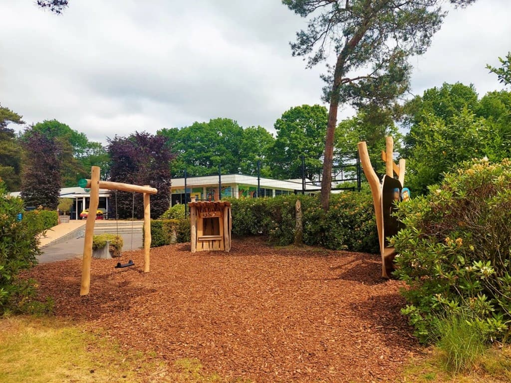Speeltuin met houten schommels en klimtoestel, omgeven door groen met het parkgebouw op de achtergrond.