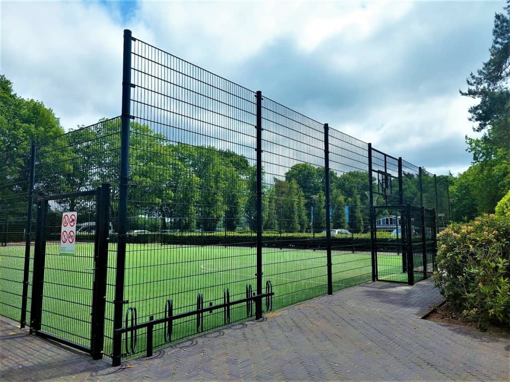 Sportveld omheind met gaas en een groene grasmat, omringd door bomen en een wandelpad in de buurt