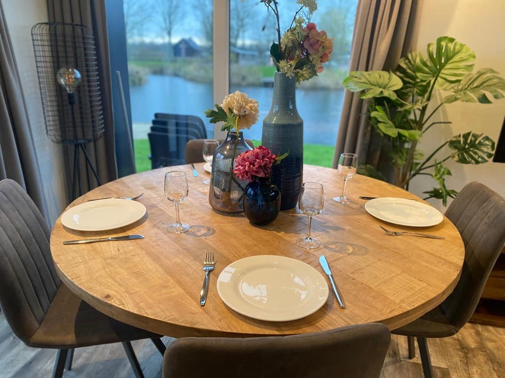 Eettafel met ronde houten blad, vier stoelen en vazen met bloemen, uitzicht op water en groen in de achtergrond