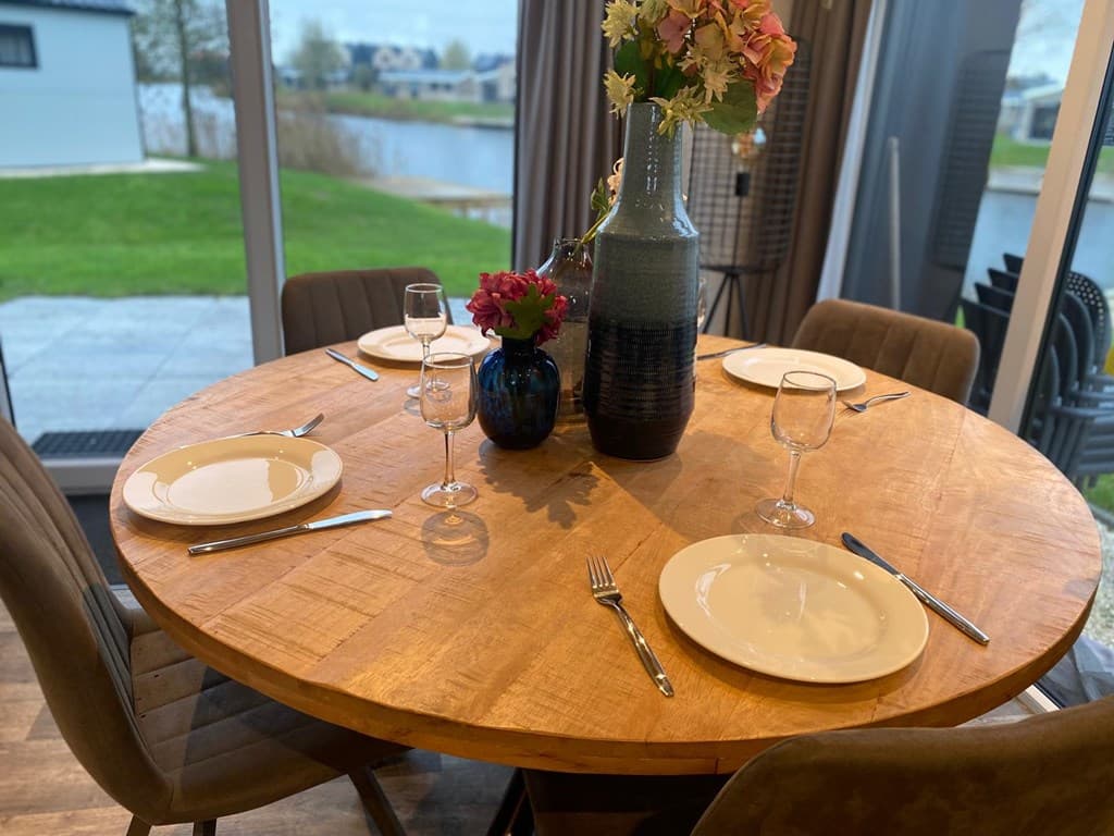 Eetkamertafel met stijlvolle borden, glazen en een vaas met bloemen, uitzicht op water en groen buiten.