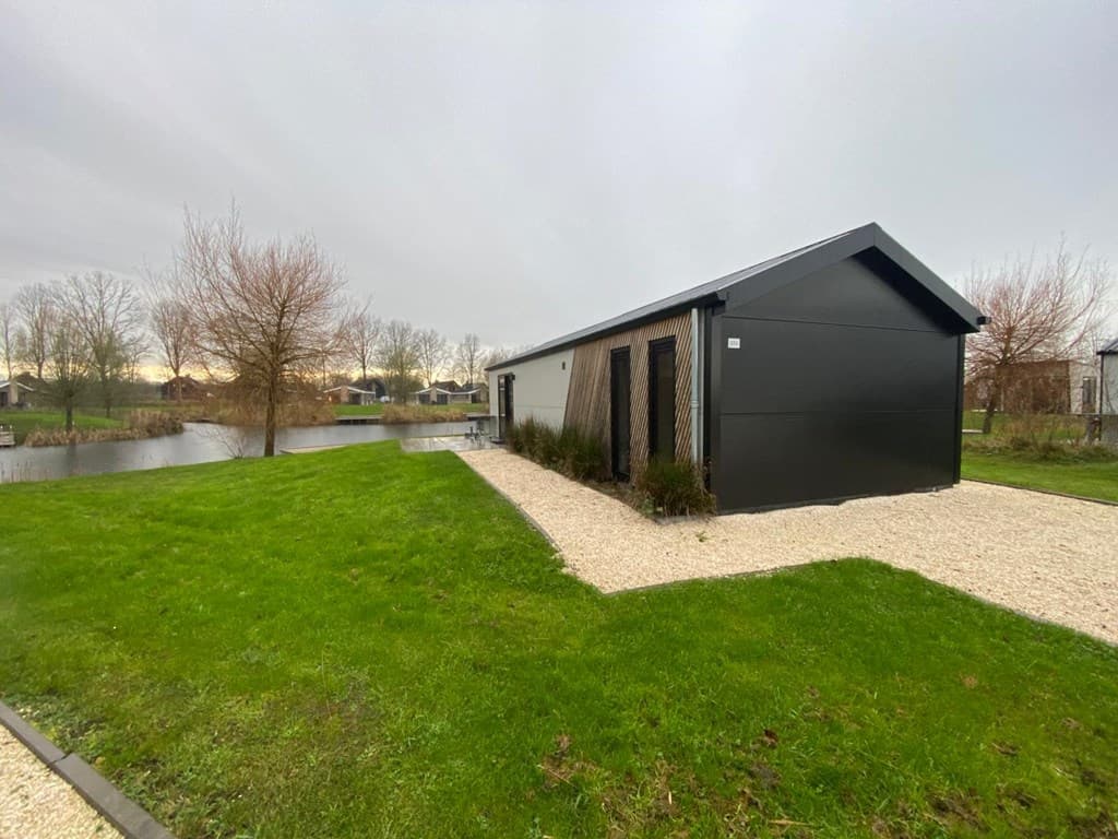 Moderne vakantiewoning aan water met een grasveld en grindpad, omringd door bomen en struiken.