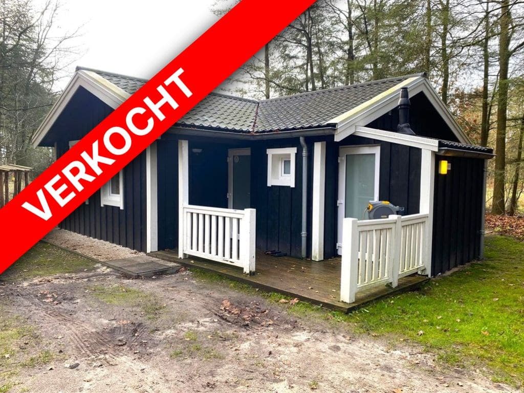 Zwarte vakantiewoning met houten veranda en twee ingangen, gelegen in een groene omgeving met begroeiing.