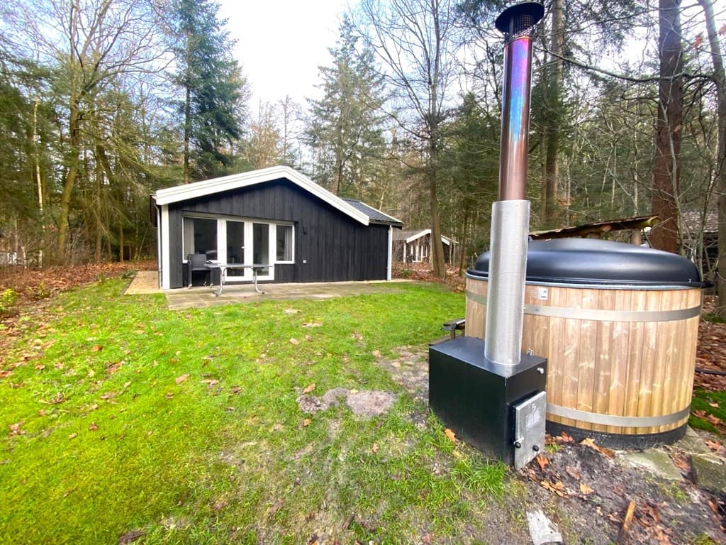 Houtachtige woning met ruime patio en vrijstaande hottub in een bosrijke omgeving met gras en bomen.