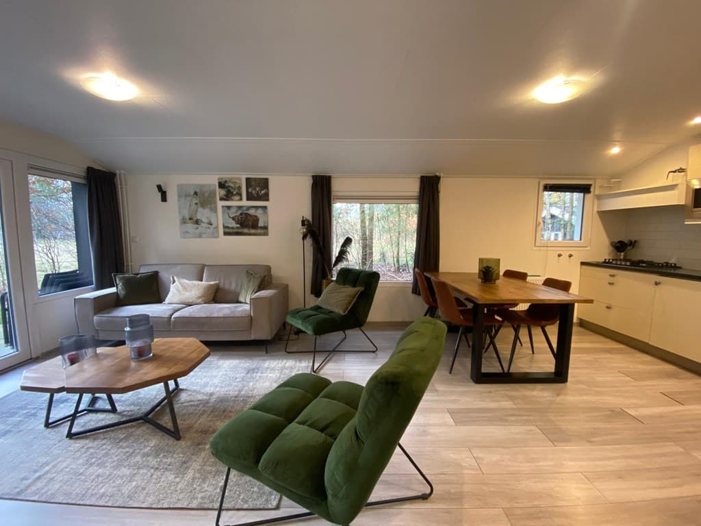 Ruime woonkamer met groene stoelen, eettafel en open keuken, grote ramen met uitzicht op de natuur