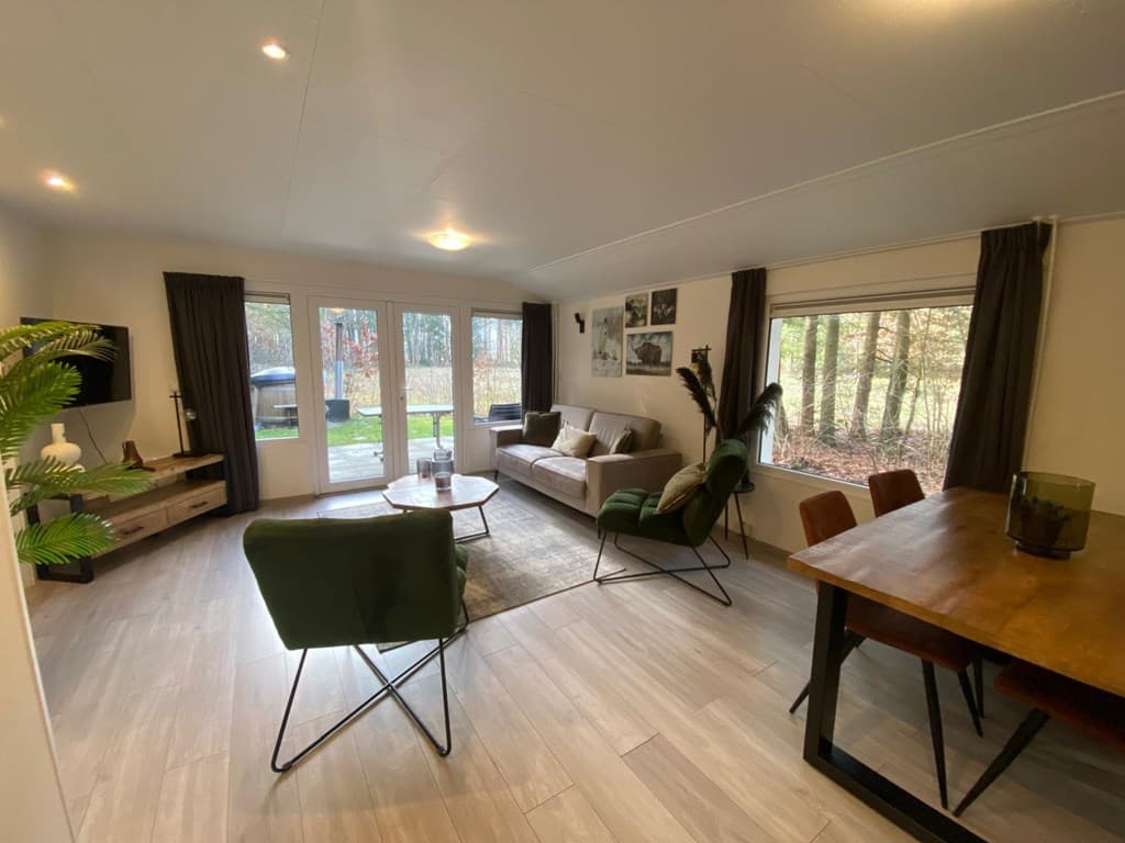 Gezellige woonkamer met banken, eettafel en grote ramen met uitzicht op de bosachtige omgeving.
