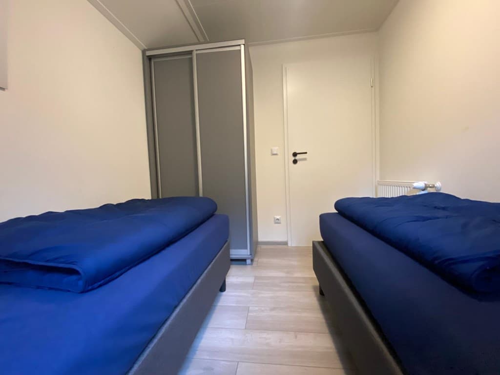 Slaapkamer met twee aparte bedden en een grijze kast aan de muur, lichte vloer en neutrale muren.